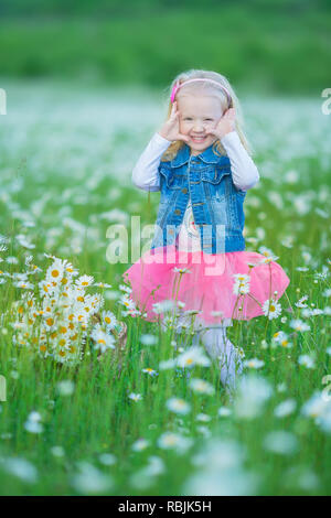 Cute little smiling baby girl en champ camomille petite blonde enfant avec couronne au chef de camomille portait un jean et la jupe rose bénéficiant d'hide-et-s Banque D'Images