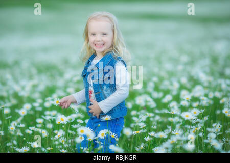 Cute little smiling baby girl en champ camomille petite blonde enfant avec couronne au chef de camomille portait un jean et la jupe rose bénéficiant d'hide-et-s Banque D'Images