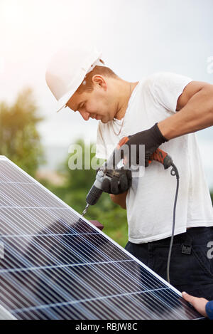 Close-up de travailleur professionnel en casque et gants de protection de la connexion du panneau photovoltaïque à la plate-forme à l'aide de tournevis électrique disponible sur Banque D'Images