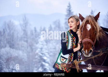 Jolie femme en blanc sans manches courtes brodées et décorées de mouton manteau de fourrure à l'extérieur à l'paire de bon cheval dans le faisceau d'hiver ensoleillée da Banque D'Images