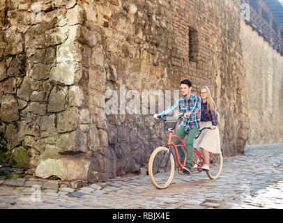 Young attractive happy tourist couple en habits, barbu et blonde woman riding on modern tandem sur la chaussée vide à l'angle Banque D'Images