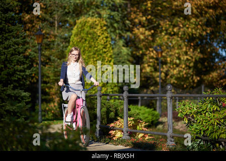 Happy blonde aux cheveux longs femme voyageur attrayant dans les verres et les vêtements modernes vélo dame rose vélo le long parc étroit pont sur bokeh backg Banque D'Images