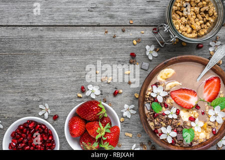 De cacao ou de chocolat et de bananes smoothie protéines bol à céréales, fraises, fleurs et graines de grenade pour le petit-déjeuner avec cuillère sur rustic Banque D'Images