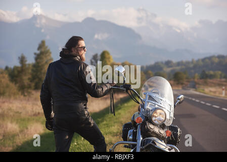 Sport motocycliste barbus dans des lunettes, des vêtements de cuir noir debout à moto cruiser moderne sur fond de paysage rural, vert Banque D'Images