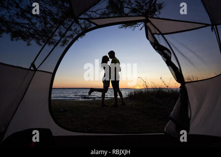 Vue de l'intérieur tente de tourisme. Silhouettes Sombres de young tourist couple romantique, man and woman standing hugging on lake shore sur bleu ciel du soir et Banque D'Images