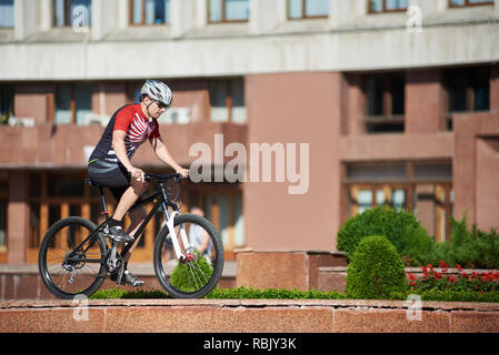 Cycliste professionnel masculin riding bike sur bordure de rue près de parterres et arbres en face du bâtiment rouge. Formation de sportif, se préparer pour concours, l'amélioration des compétences. Concept de vie sain Banque D'Images