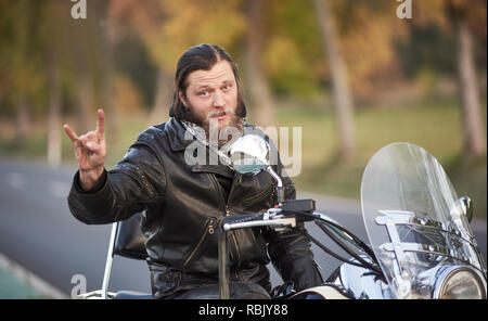 Beau jeune motard barbu en vêtements de cuir noir assis sur moto puissante moderne brillant montrant cornes signe sur l'arrière-plan flou flou vintage de golden arbres sur journée ensoleillée. Banque D'Images