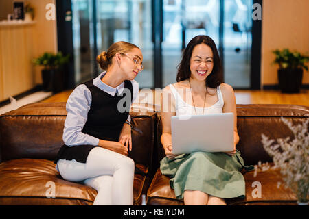 Deux collègues dans un milieu de travail diversifié ont une réunion ensemble sur un canapé dans un bureau à la mode. Une femme est de race blanche et les autres pays asiatiques Banque D'Images