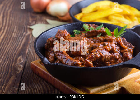 Bœuf Stroganoff à la sauce tomate et la crème sure et de pommes de terre frits servis dans la poêle en fonte sur table en bois rustique Banque D'Images