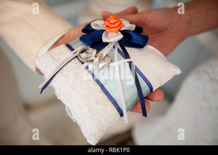 Une paire de bagues de mariage, entrelacées avec ruban blanc et bleu sur l'oreiller. L'oreiller est dans les paumes de la mariée et le marié. Banque D'Images