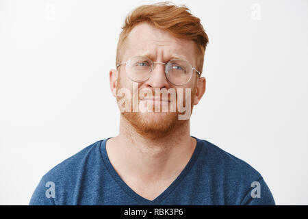 Beau mâle roux sombre avec barbe et sourcils lèvres pinçant à droite avec regret et déception, debout sur fond gris dans des vêtements décontractés et verres Banque D'Images