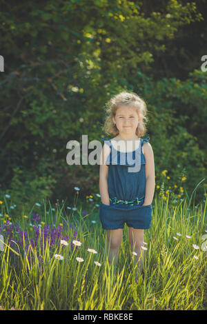 Cute blonde jeune fille élégante de l'enfant habillé de bleu jeans dress posing sur prairie de camomille sauvage forêt.scène Adoreable. Banque D'Images