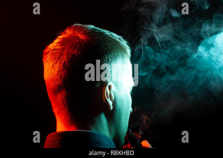 Vue de l'arrière sur la tête d'un homme dans une chemise de fumer la vape et exhalant la fumée colorée de vert et rouge à différentes directions de lui Banque D'Images