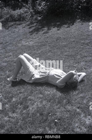 Années 1950, l'heure d'été et d'une jeune femme portant une robe de coton léger allongé sur le sol avec son bras à travers une partie de son visage... comme pour dire vous pouvez me photo mais sais pas moi ! Banque D'Images