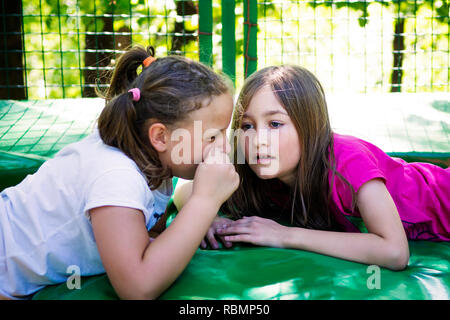 Deux filles de dire un secret, allongé sur le trampoline, piscine dans parc verdoyant. Banque D'Images