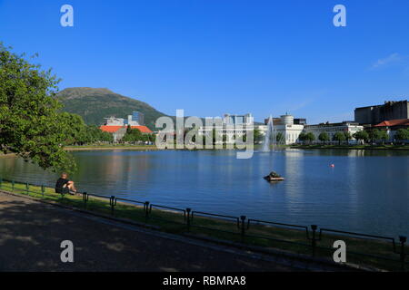 Un homme est assis sur la rive du lac, Lille Lungegårdsvannet, dans la ville de Bergen, en Norvège, avec une vue sur le mont Ulriken, KODE 4 et d'autres bâtiments. Banque D'Images