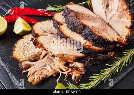 Close-up de jambon de porc rôti sur une pierre noire plateau avec le romarin, l'ail et les tranches de lime, horizontal Vue de dessus Banque D'Images