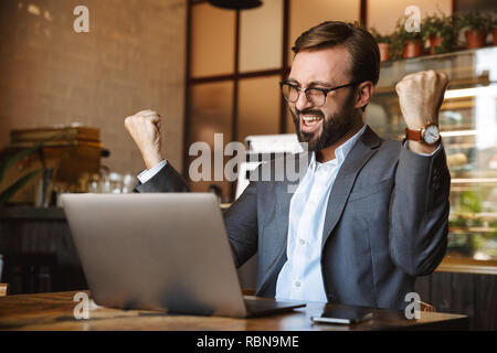 Beau jeune homme habillé en fonction de travailler sur un ordinateur portable, assis au café à l'intérieur, des succès à célébrer Banque D'Images