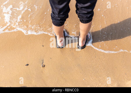 Les jambes de la jeune femme debout sur une plage de sable avec des vagues déferlaient sur ses pieds Banque D'Images