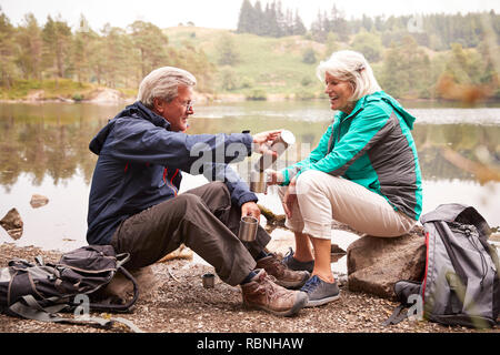 Senior couple sitting by a lake pouring coffee d'une fiole pendant un camping holiday, Lake District, UK Banque D'Images