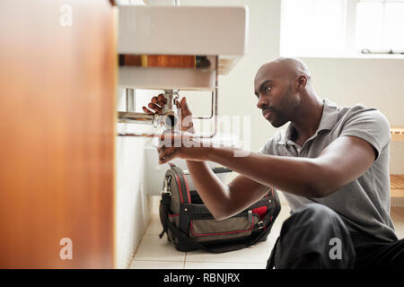 Plombier jeune homme noir assis sur le plancher la fixation d'un évier de salle de bains, vue de la porte Banque D'Images