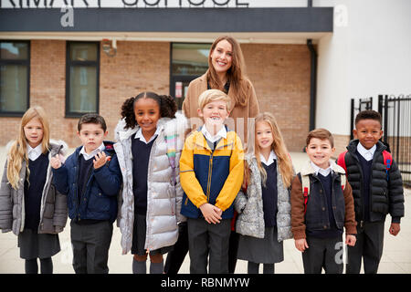 Les enfants de l'école primaire en face de l'école avec leur enseignant à la recherche d'appareil photo, vue de face Banque D'Images