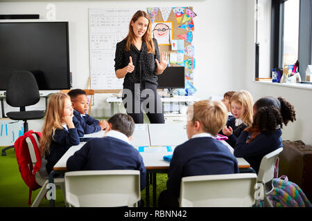 Enseignant de l'école des femmes debout dans une salle de classe aux élèves des gestes, assis à une table à l'écoute Banque D'Images