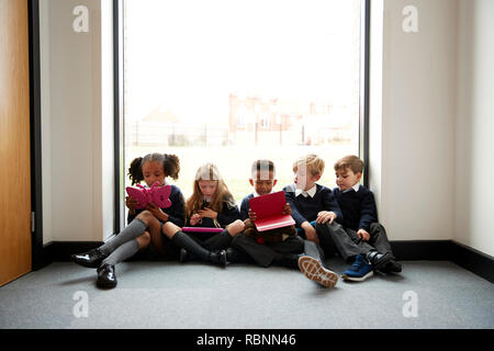 L'école primaire les enfants assis dans une rangée sur le sol en face d'une fenêtre dans un couloir d'école à l'aide d'ordinateurs tablettes, front view Banque D'Images