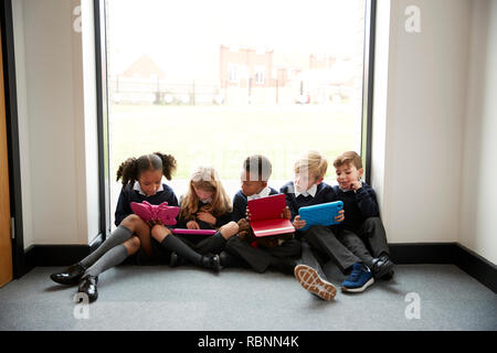 L'école primaire de cinq enfants assis dans une rangée sur le sol en face d'une fenêtre dans un couloir d'école à la tablette à l'ordinateur, vue de face, Close up Banque D'Images