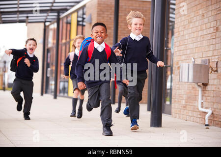 Heureux les enfants de l'école primaire, le port d'uniformes scolaires et de sacs à dos, s'exécutant sur une passerelle à l'extérieur de leur bâtiment scolaire, vue de face, Close up Banque D'Images