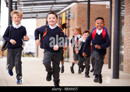 L'école primaire les enfants excités, le port d'uniformes scolaires et de sacs à dos, s'exécutant sur une passerelle à l'extérieur de leur bâtiment scolaire, vue de face, Close up Banque D'Images