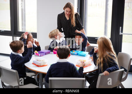 Enseignante est de parler à un groupe d'école primaire enfants assis ensemble à une table ronde à manger leurs paniers-repas Banque D'Images