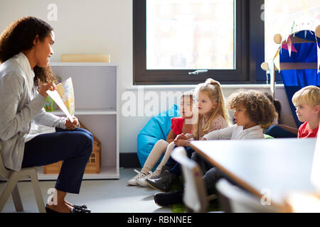 Vue latérale d'un enseignant de maternelle femme assise sur une chaise montrant un livre pour enfants dans une salle de classe Banque D'Images