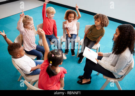 Jeune femme professeur de l'école de la lecture d'un livre à l'enfant Les enfants de l'école, assis sur des chaises en cercle dans la salle de classe la main pour répondre à une question, elevated view Banque D'Images