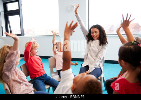 Une classe de l'école infantile des enfants assis sur les chaises en cercle dans la salle de classe, soulevant les mains avec leur enseignante, Close up Banque D'Images