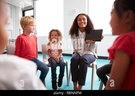 Plus d'épaule de souriante jeune femme professeur de l'école montrant un ordinateur tablette à l'enfant les enfants, assis en cercle dans la salle de classe, plus d'épaule Banque D'Images