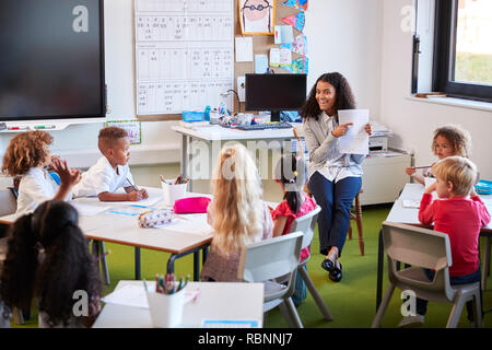 Female infant school teacher assis sur une chaise en face d'enfants de l'école dans une classe holding up et d'expliquer une feuille de travail pour eux Banque D'Images