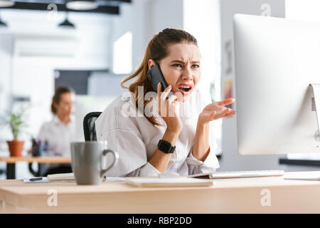 Choqué déçu young businesswoman habillé en shirt assise à son lieu de travail au bureau, talking on mobile phone Banque D'Images