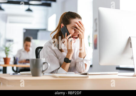 Choqué déçu young businesswoman habillé en shirt assise à son lieu de travail au bureau, talking on mobile phone Banque D'Images
