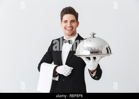 Happy young waiter in tuxedo holding plateau avec cloche métallique et une serviette Banque D'Images