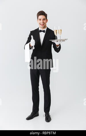 Full Length portrait of a smiling young waiter in tuxedo holding tray with deux verres de champagne sur fond blanc Banque D'Images