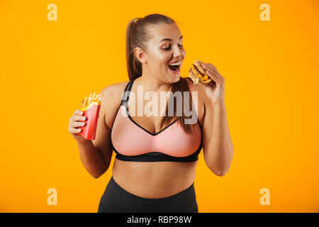 Portrait d'un embonpoint heureux jeune femme portant des vêtements de sport isolés sur fond jaune, manger burger et frites Banque D'Images