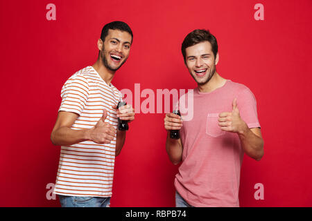 Deux jeunes hommes gais isolés sur fond rouge, de boire l'eau de soude à partir de bouteilles, Thumbs up Banque D'Images