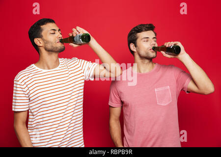 Deux jeunes hommes gais isolés sur fond rouge, de boire l'eau de soude à partir de bouteilles Banque D'Images