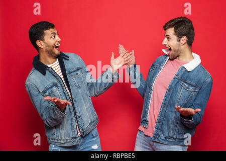 Deux jeunes hommes excités amis portant des vestes en denim isolés sur fond rouge, la célébration de Banque D'Images