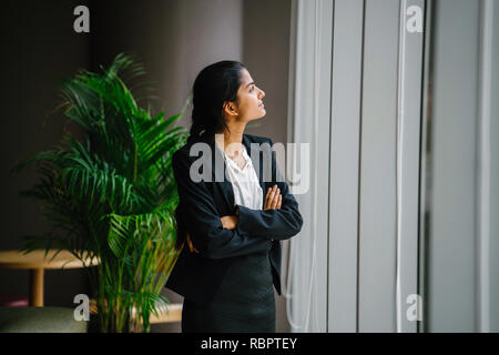 Portrait d'une jeune et jolie femme asiatique indien dans un costume standing par une fenêtre au cours de la journée. Elle sourit alors qu'elle contemple la vue. Banque D'Images