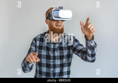 Cheveux rouge mature man wearing casque de réalité virtuelle isolés sur mur gris pointant au space regardant la vidéo close-up Banque D'Images