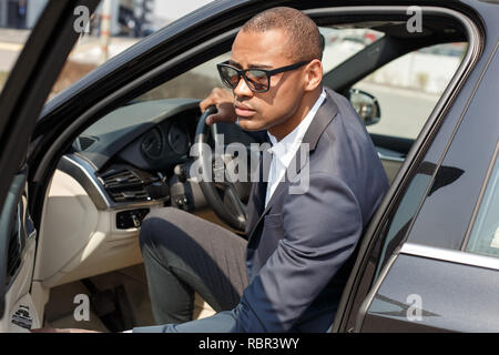 Jeune homme mulâtre portant des lunettes de soleil dans la voiture porte ouverte maintenant le volant à côté des Banque D'Images