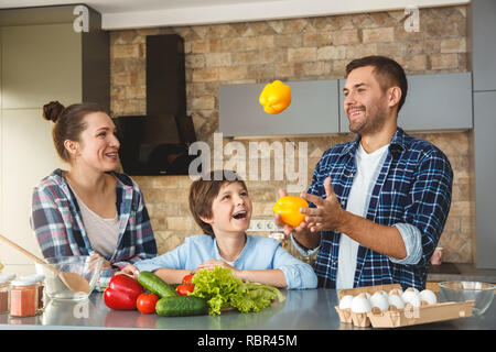 Mère Père et fils à la maison debout dans la cuisine ensemble woman and boy looking at man juggling joyeux sourire excité poivrons Banque D'Images