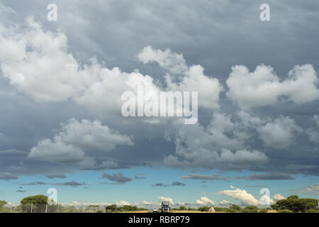 Cumulus ou puffy nuages dans le ciel et au-dessous d'une longue route qui s'étend à l'infini avec des véhicules dans la province du nord-ouest de l'Afrique du Sud Banque D'Images